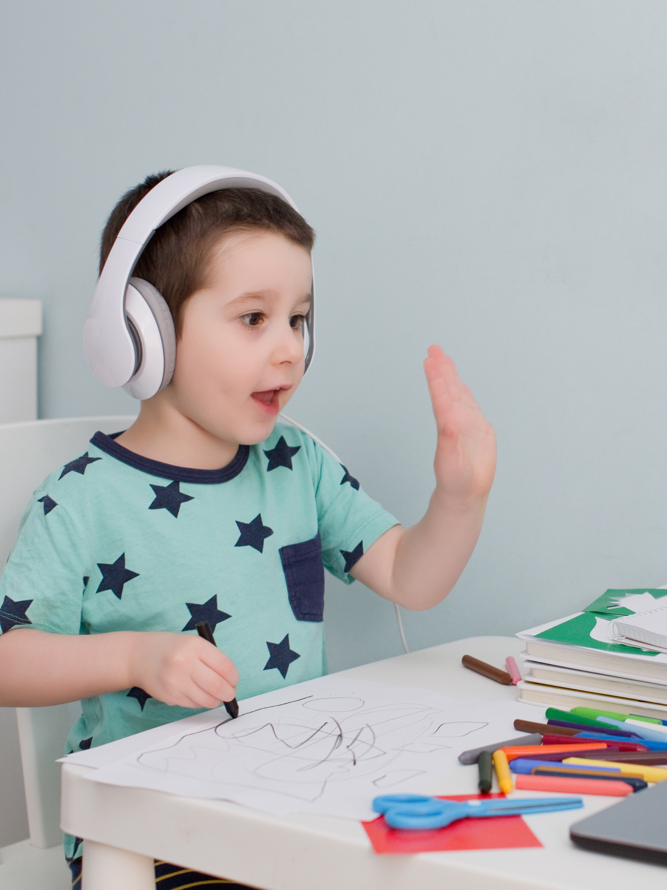 A young boy wearing headphones sitting at a table, drawing with markers on a sheet of paper, and raising one hand.