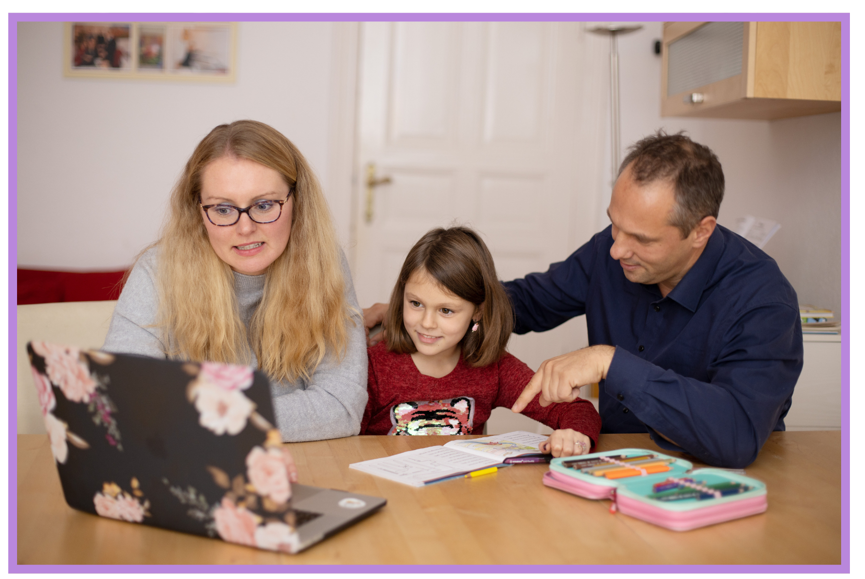 Family of three, including a woman, a young girl, and a man, gathered at a table looking at a laptop with school supplies and open notebooks. They are learning together.