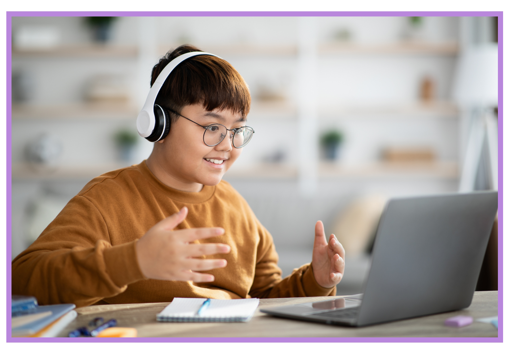 A young person with glasses and short brown hair wears white headphones and smiles while engaging in a video call or online class on a laptop. They sit at a table with notebooks, pens, and office supplies, in a bright, modern room.
