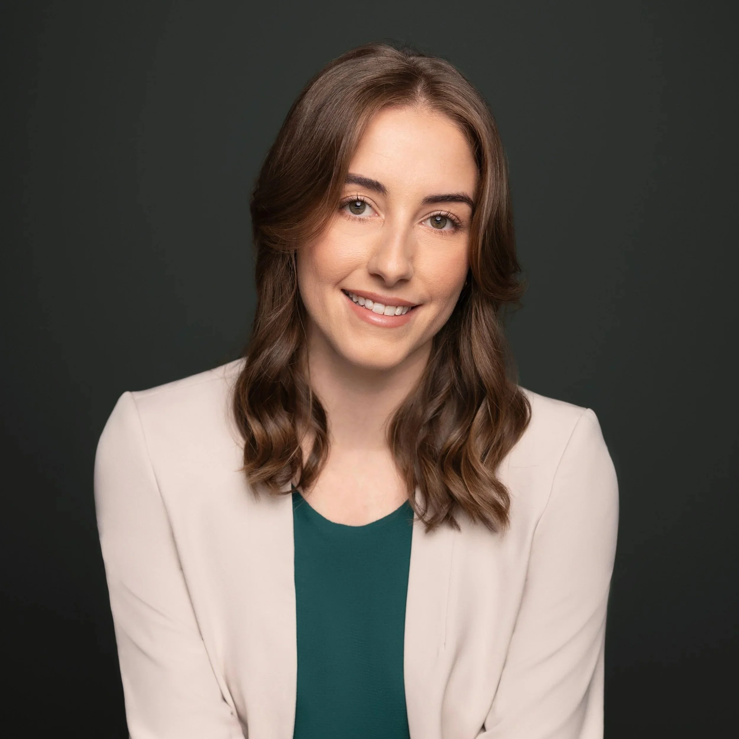 Headshot of a woman with brown hair styled in loose waves, wearing a beige blazer over a dark green top, smiling against a dark gray background.