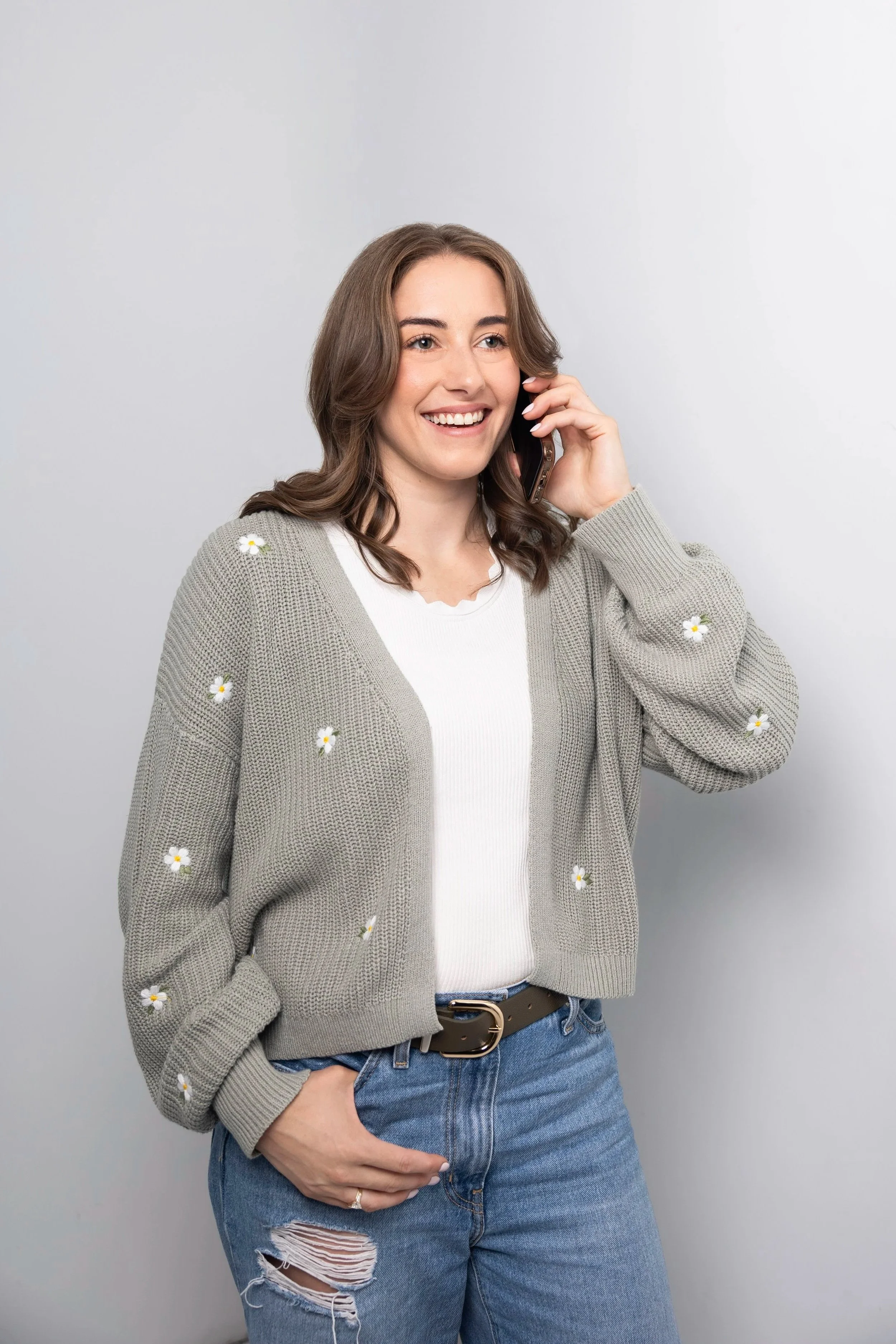 A young woman with brown hair and a bright smile talking on her cellphone. She is wearing a beige cardigan with small floral patterns, a white top, and ripped blue jeans with a belt, standing against a plain light gray background.