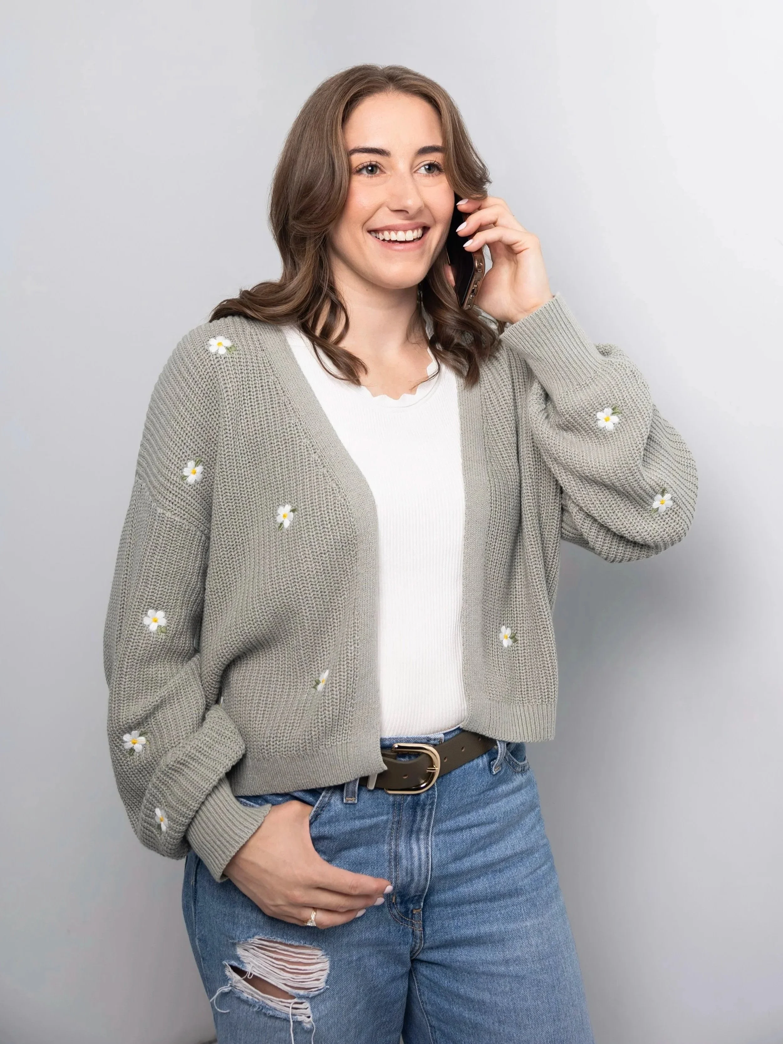 A young woman with brown hair, smiling, talking on a cellphone, wearing a light gray cardigan with small daisy patterns, a white shirt, blue ripped jeans, and a black belt, standing against a plain gray background.