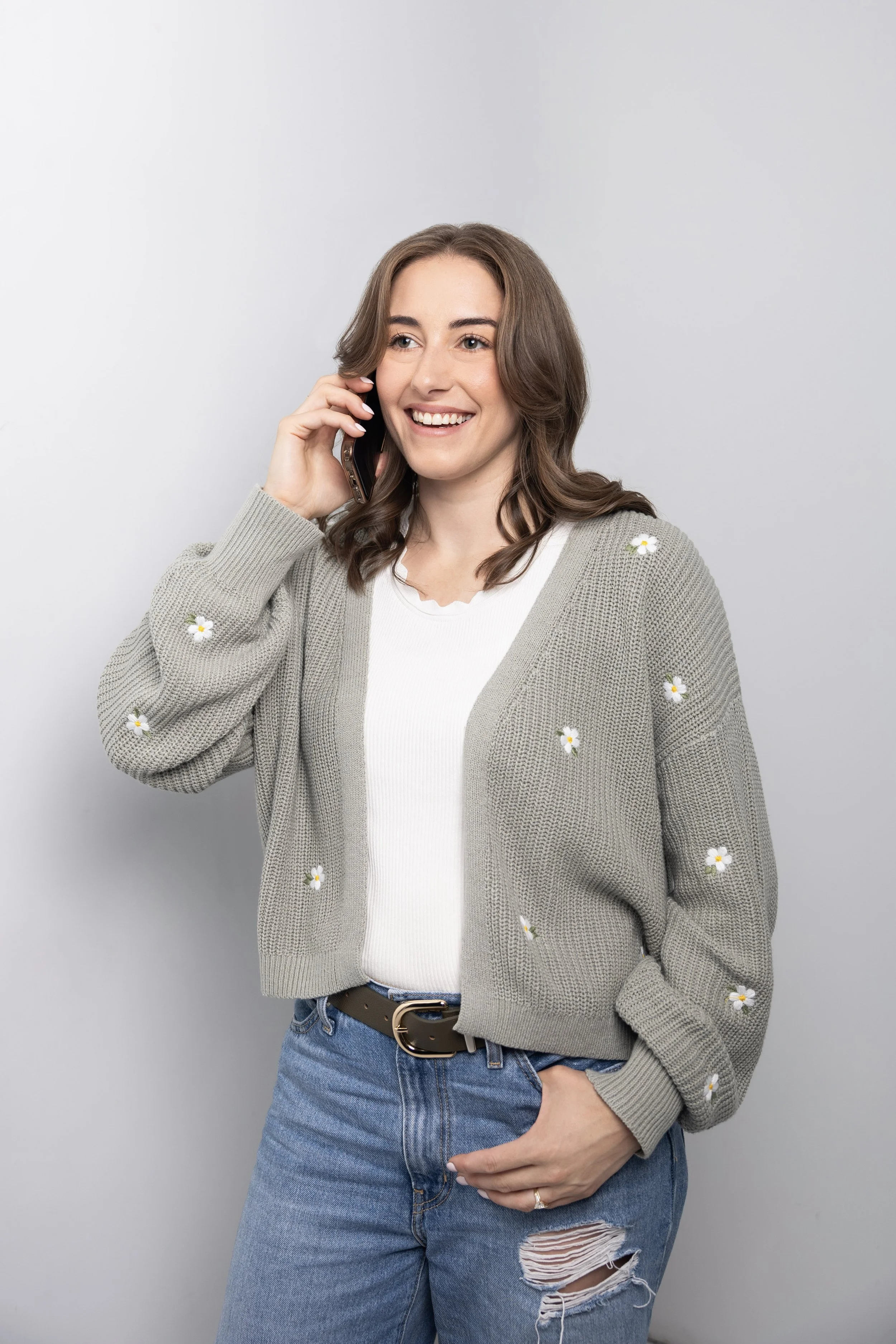 A young woman with shoulder-length brown hair, smiling, holding a cell phone to her ear. She is wearing a beige cardigan with daisy flower patterns, a white top, and ripped blue jeans. She is standing in front of a plain light gray background.