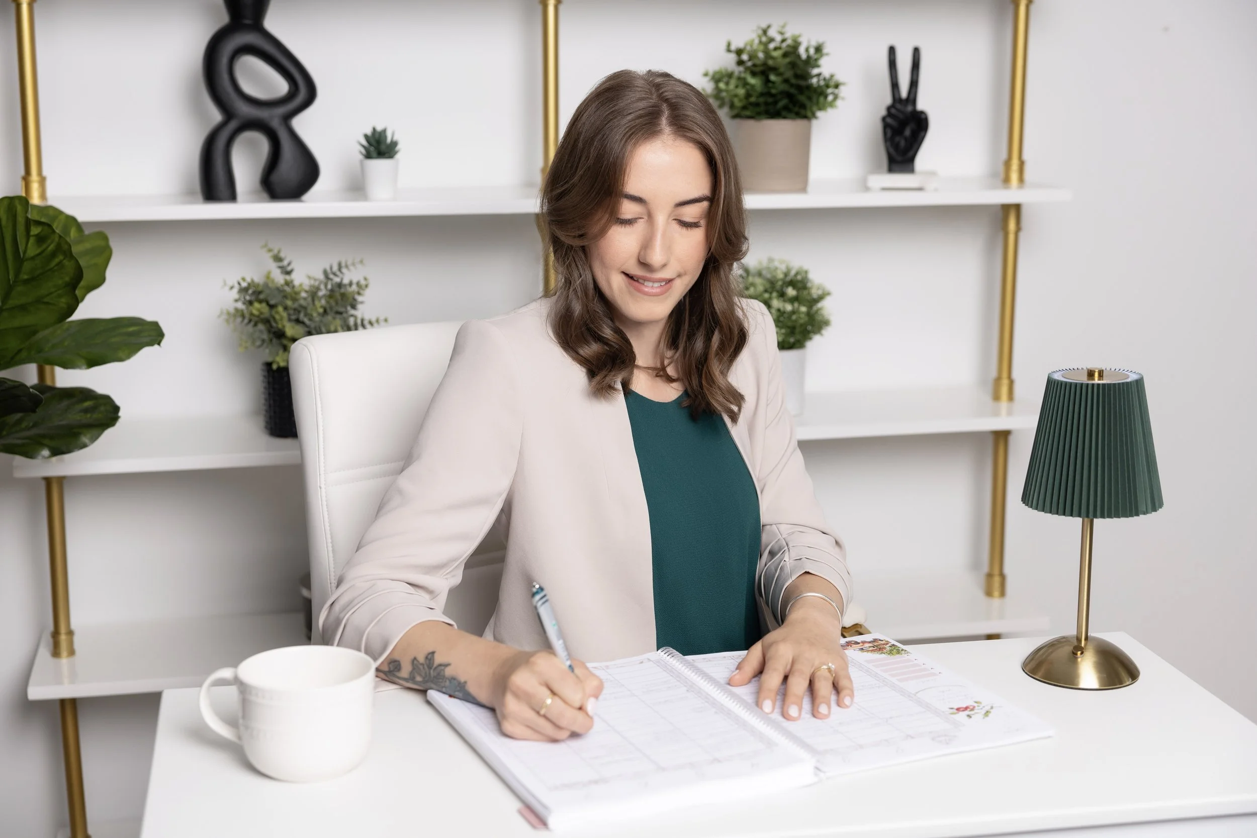 A woman with brown hair, wearing a beige blazer and teal blouse, sitting at a white desk, writing in a planner with a pen, surrounded by plants and decorative items in a modern office.