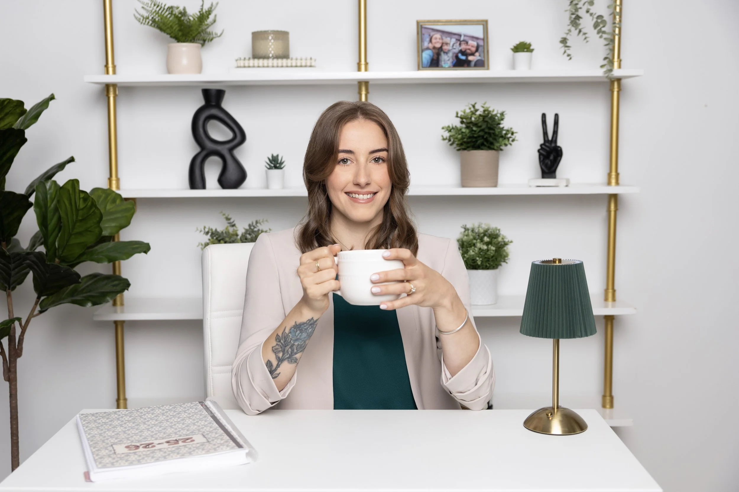 A woman with brown hair, smiling, sitting at a white desk holding a white mug. She has a tattoo on her right forearm. The background features a white wall with shelves decorated with plants, sculptures, and framed photos. There is a green table lamp on the desk.