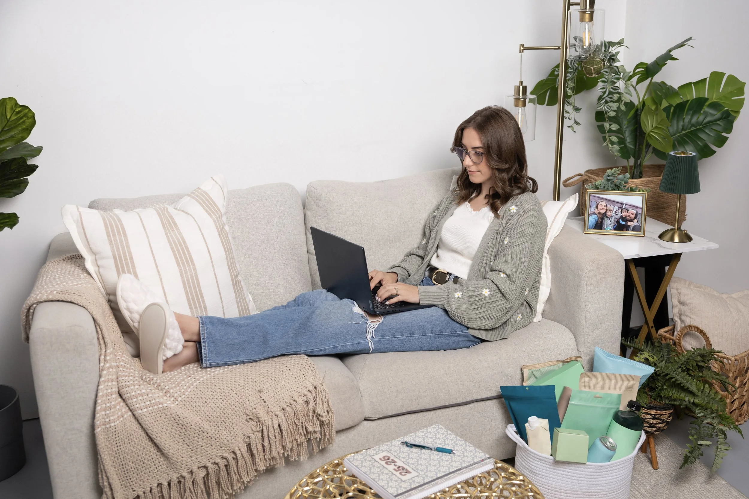 A woman with glasses and brown hair relaxing on a beige sofa with a laptop, surrounded by houseplants, a framed family photo, and various shopping bags in a well-decorated living room.