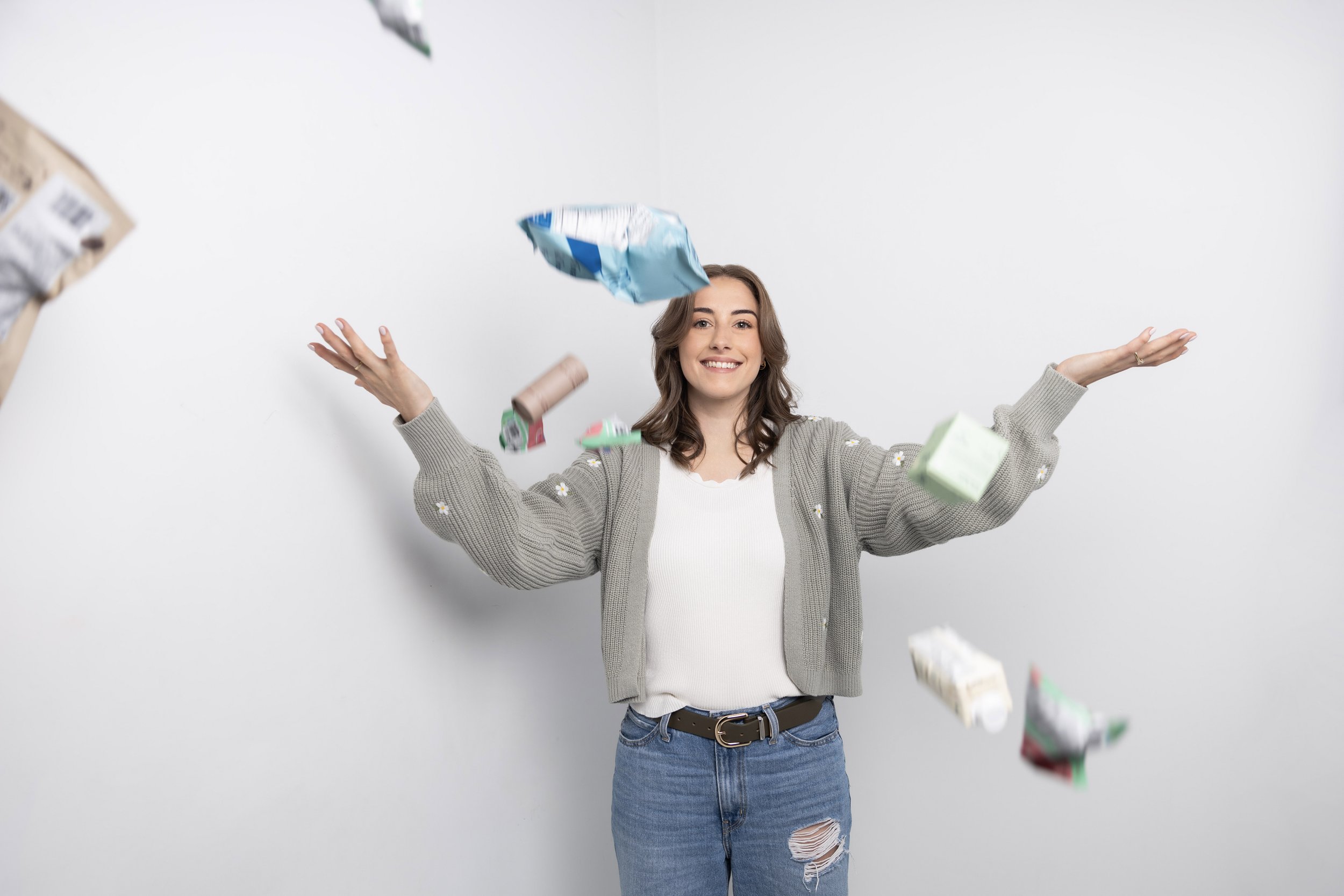 A smiling young woman in a beige cardigan and ripped jeans is throwing pieces of paper or packaging into the air against a plain white wall.