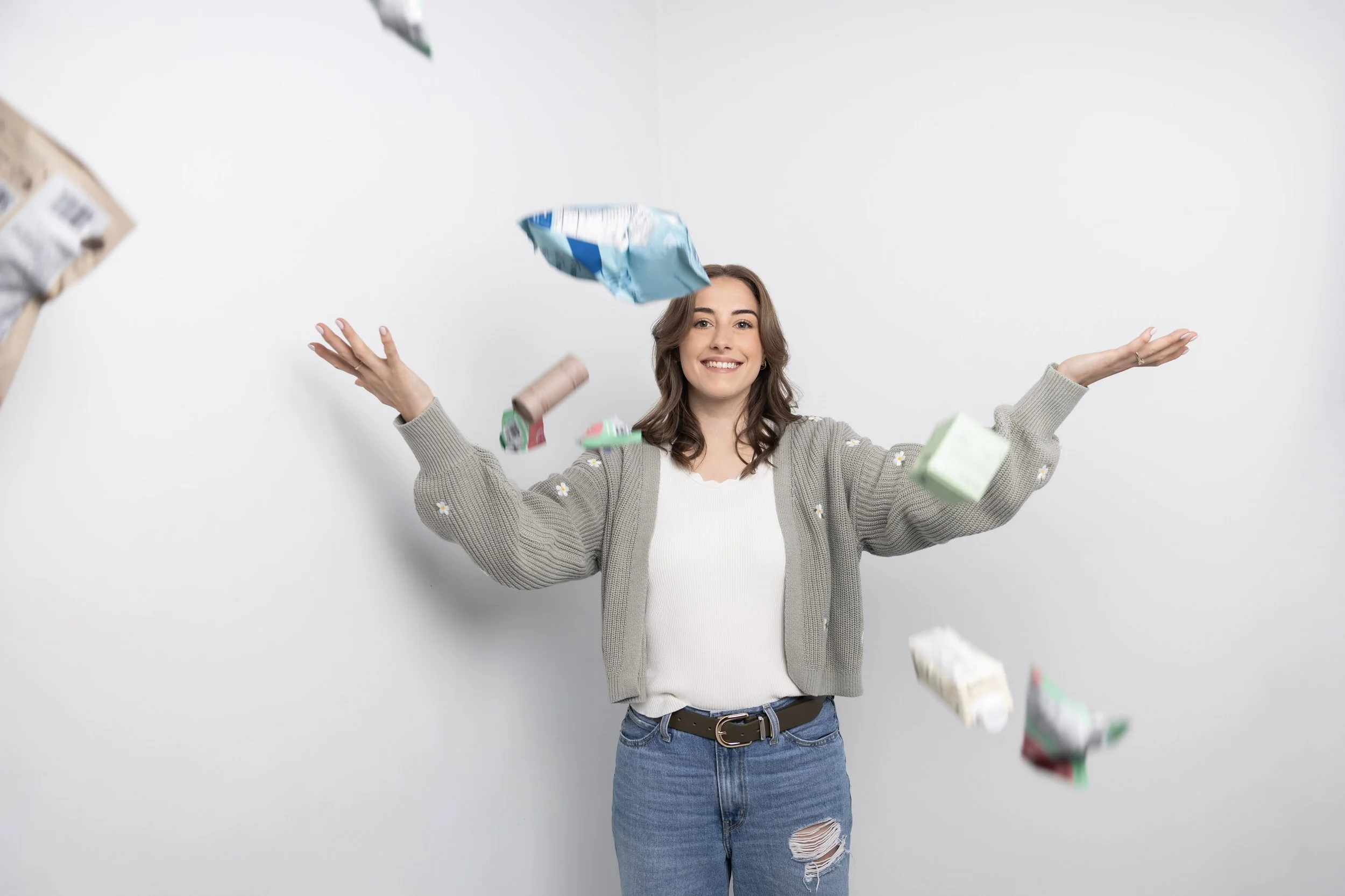 A young woman with brown hair in a gray cardigan over a white shirt and ripped jeans smiling while tossing small packages into the air in front of a plain white background.