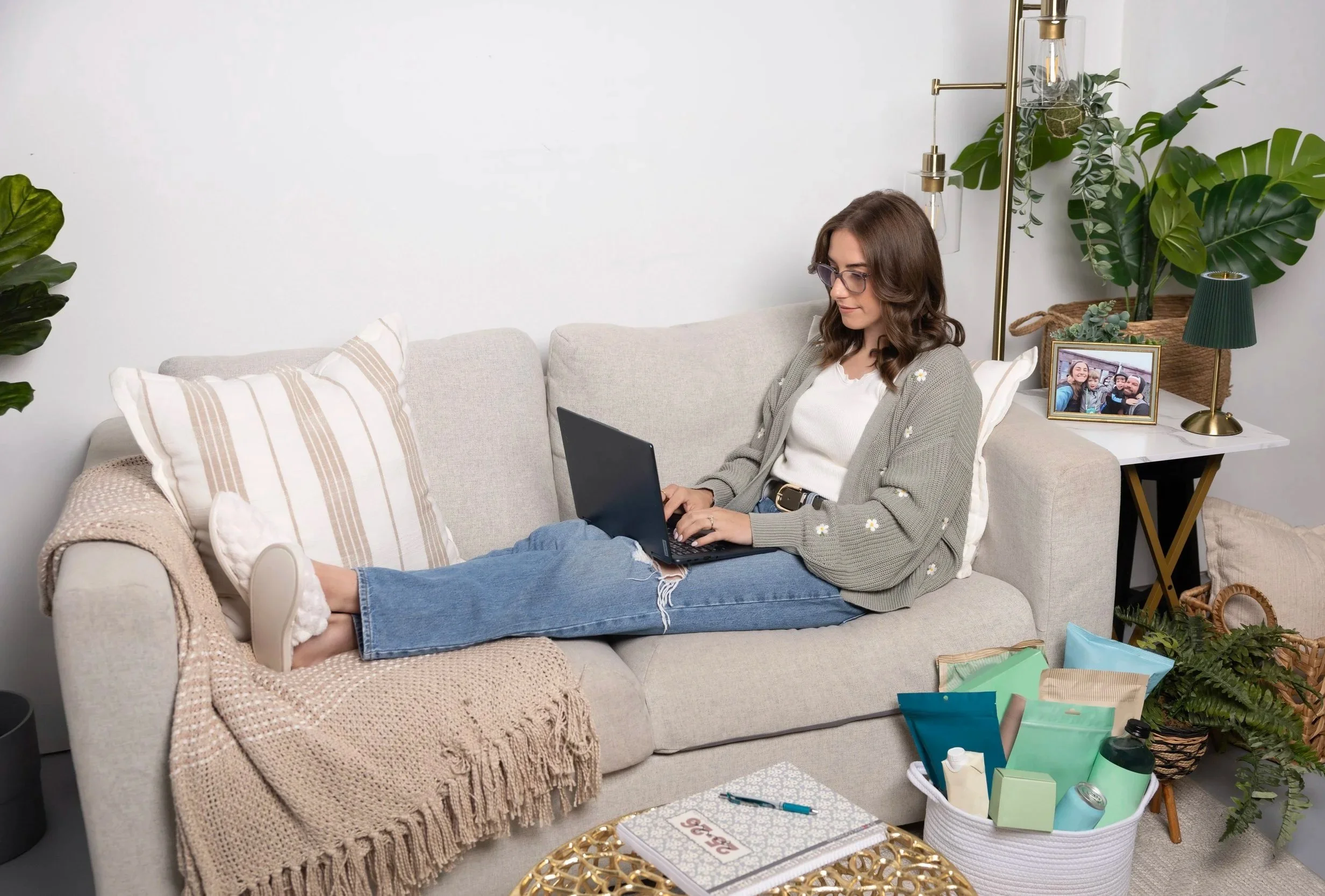 Young woman with glasses sitting on a beige couch, working on a silver laptop. She is wearing a white shirt and jeans, with a cardigan. The couch has striped pillows and a knit blanket. Next to her is a table with a framed photo, a small green lamp, and a plant. In the foreground, there are gift bags and a tray with books and items, and colorful plants and decor in the background.