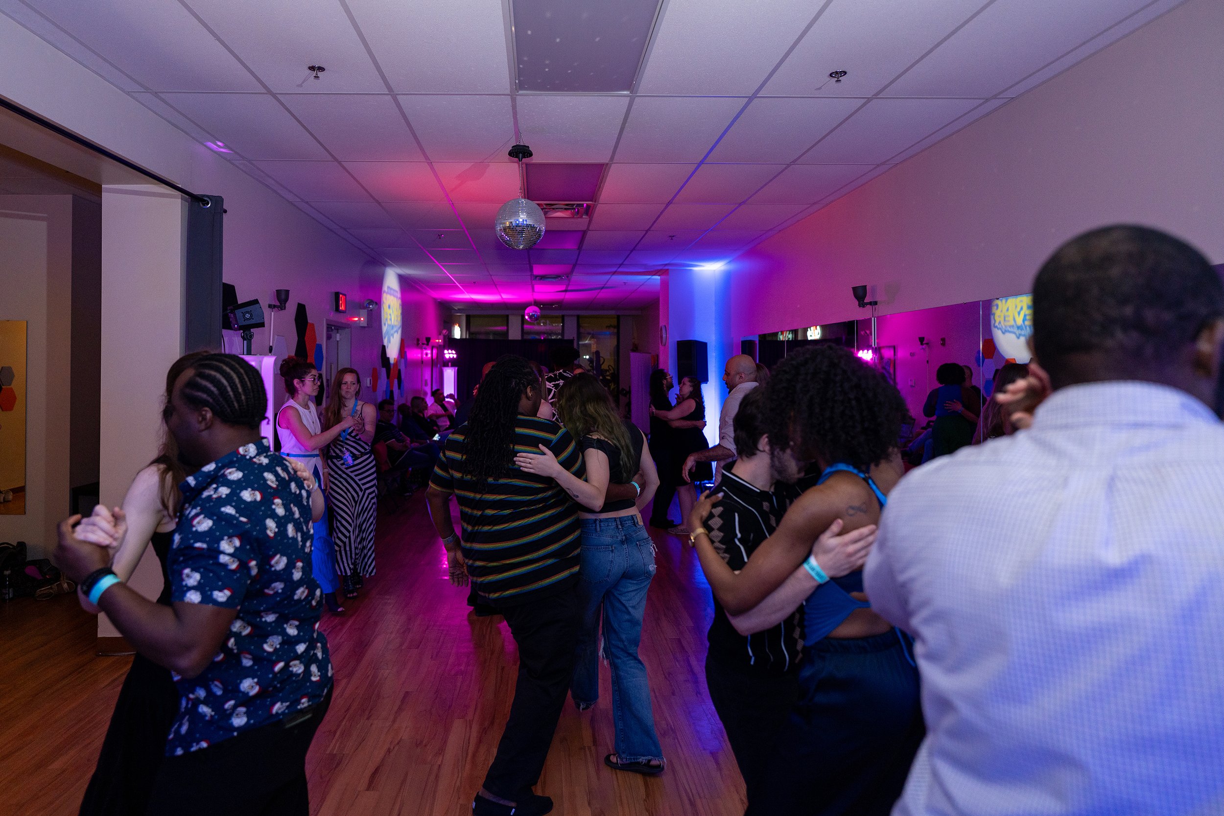 People dancing and socializing in a dimly lit room with purple and pink lighting, and a disco ball hanging from the ceiling.