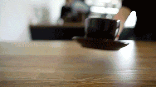 A close-up of a coffee cup on a saucer on a wooden table, with a person in the background.