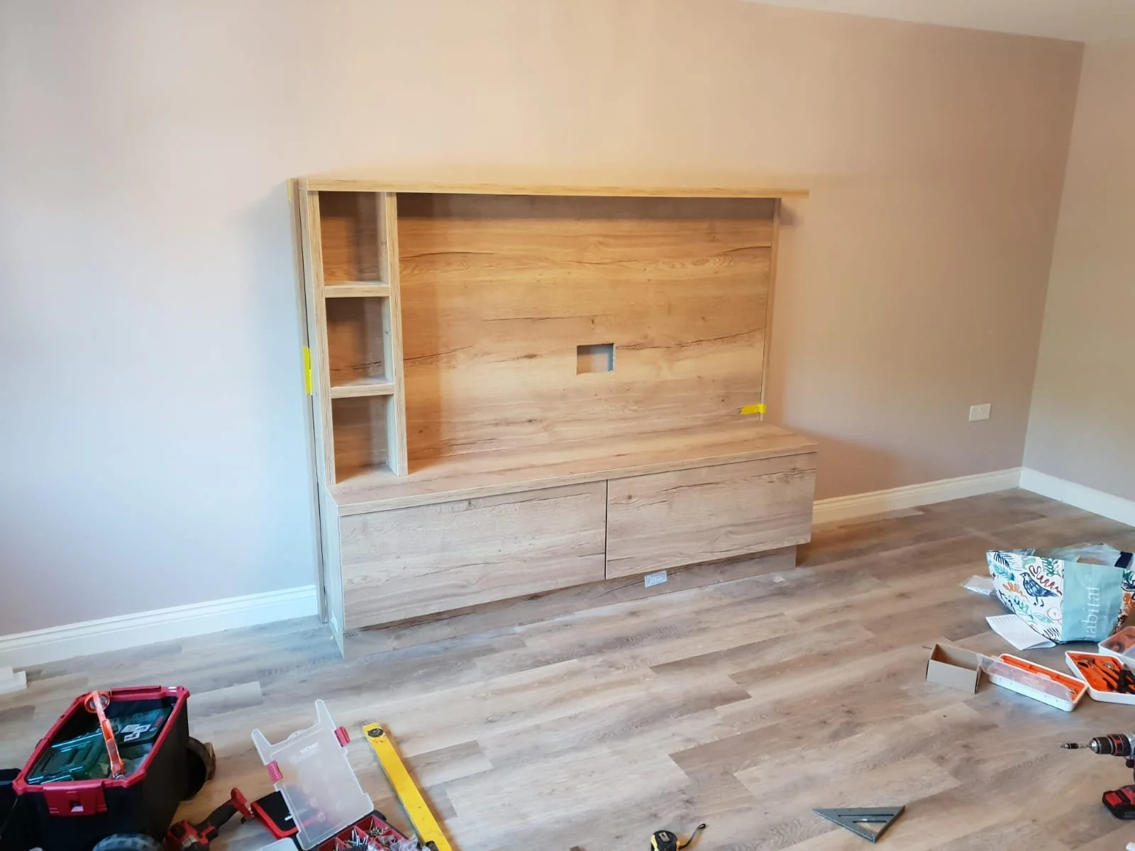Room under renovation with a partially assembled wooden entertainment center on a beige wall. Tools and supplies are scattered on the floor, including a toolbox, drill, level, and boxes of hardware.