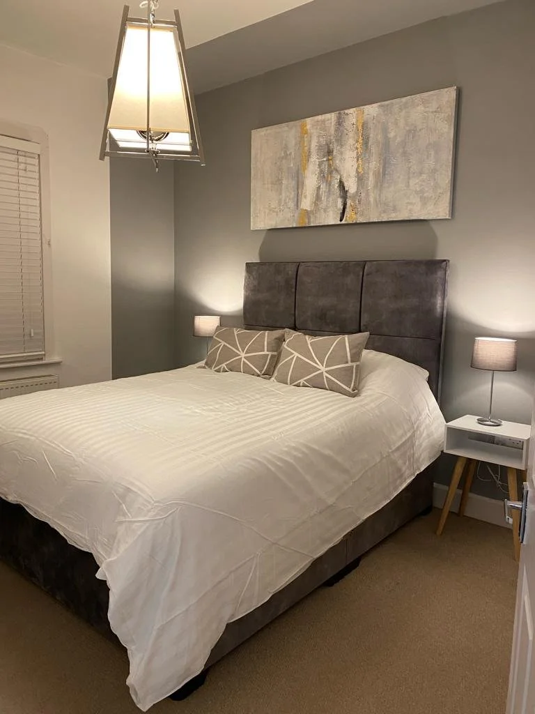 A bedroom with a bed, two nightstands with lamps, a gray upholstered headboard, a painting above the bed, and a ceiling light fixture.
