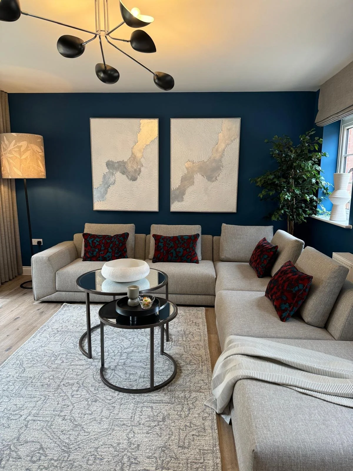 Living room with a beige sectional sofa, black coffee tables, red and black pillows, a blue accent wall, abstract artwork, a tall floor lamp, and a large potted plant near a window.