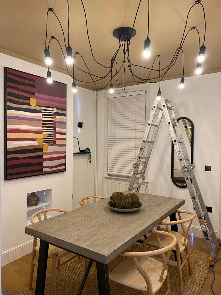 Interior of a dining room with a wooden table, six light-colored wooden chairs, and a ladder leaning against the wall near a window with closed blinds. Modern lighting fixtures hang from the ceiling, and a large abstract painting decorates the wall.