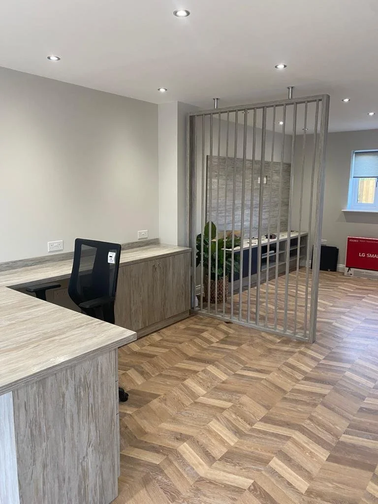 Office space with a wooden desk, an office chair, a partition with vertical bars, and a plant in a pot. There are ceiling lights, a window with blinds, and a red LG Smart TV in the background.