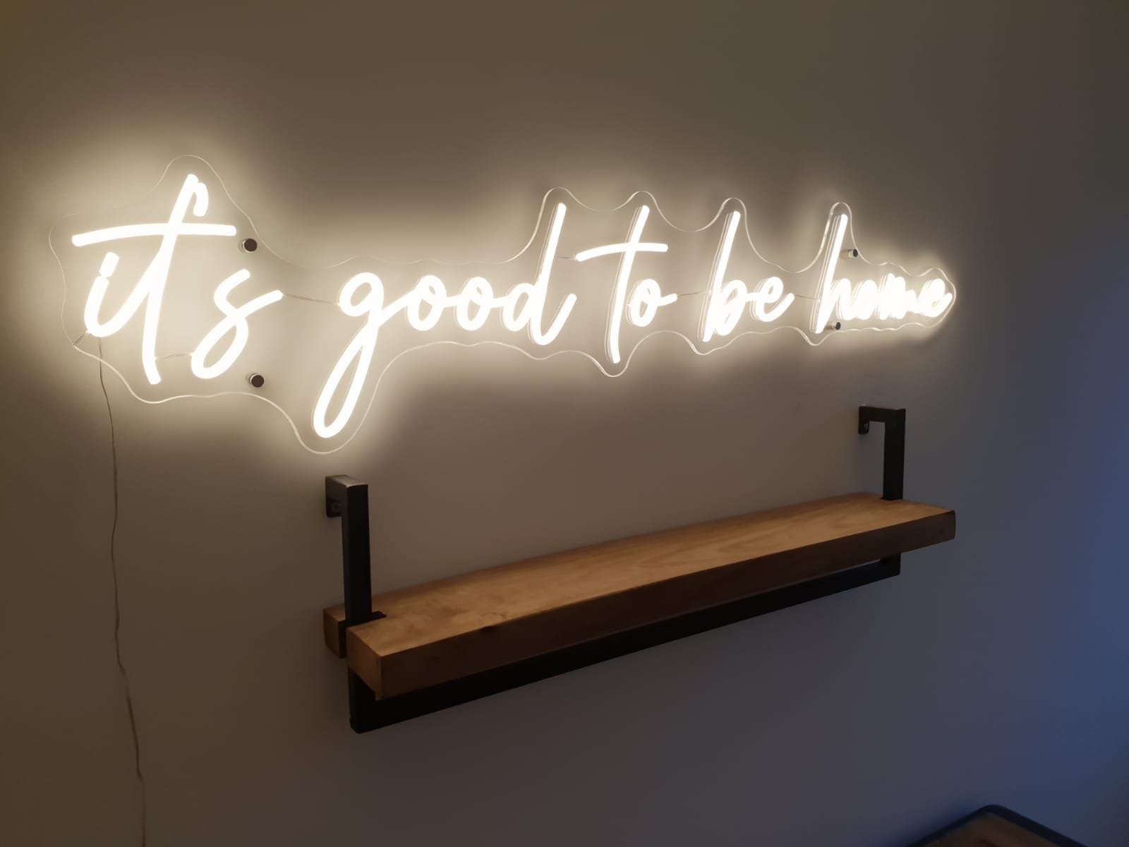 Neon sign on a wall that reads 'it's good to be home' in cursive white letters, with a wooden shelf mounted below it.