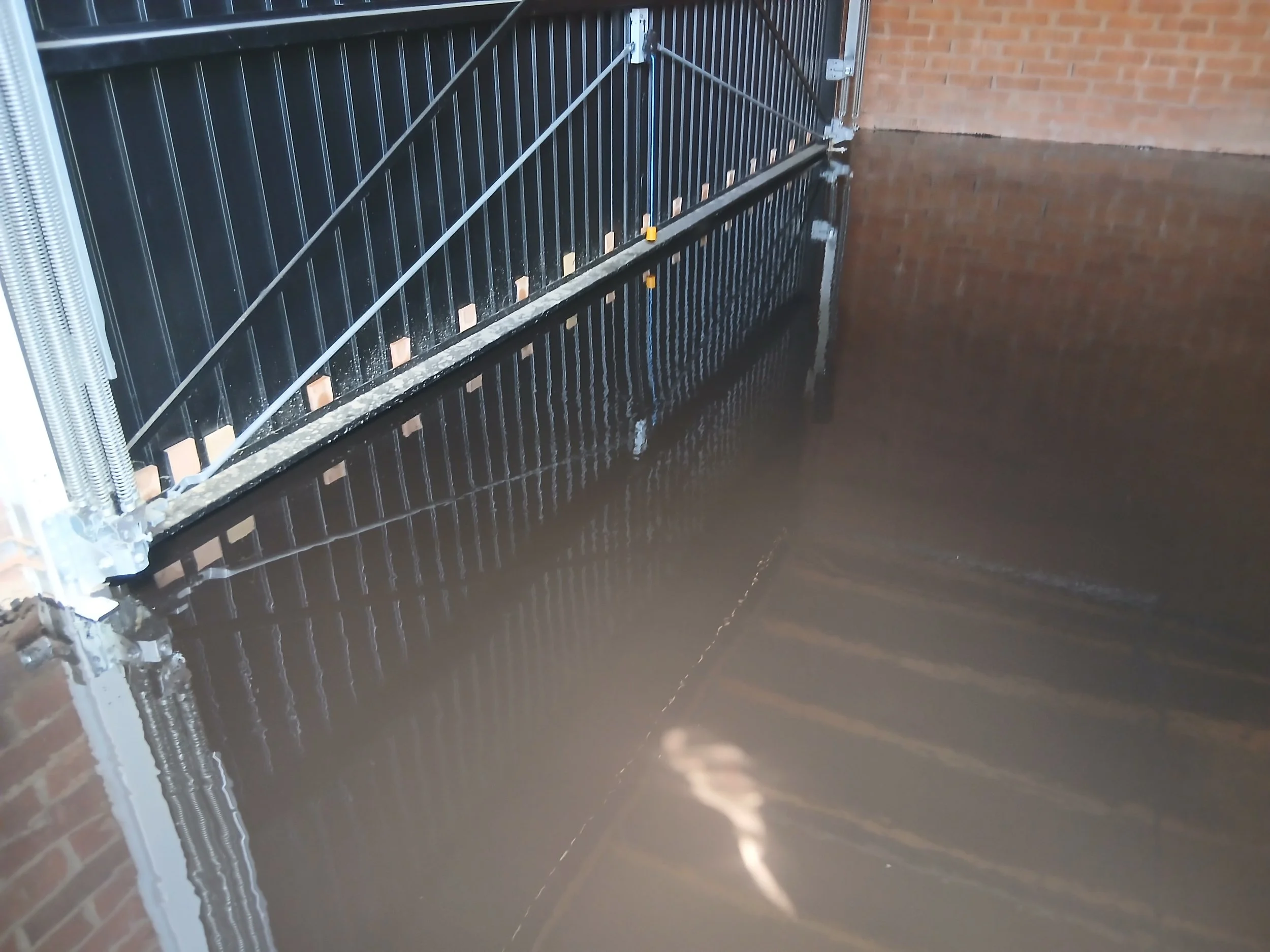 A sliding black metal gate with security bars, attached to a brick wall, with water or flood at the bottom.