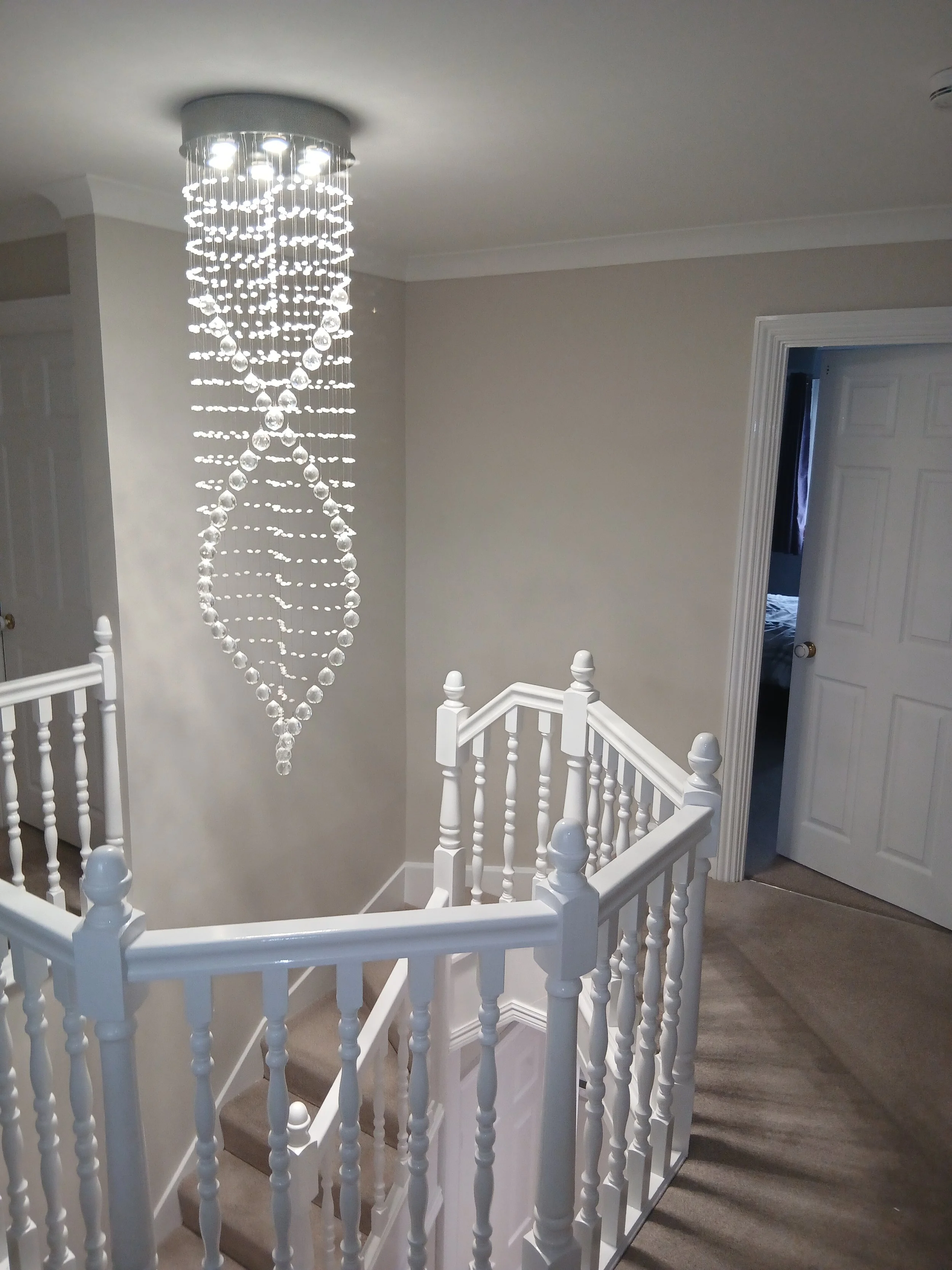 Interior view of a staircase with white wooden railing, a chandelier hanging from the ceiling, beige carpeted stairs, and an open door leading to a room.