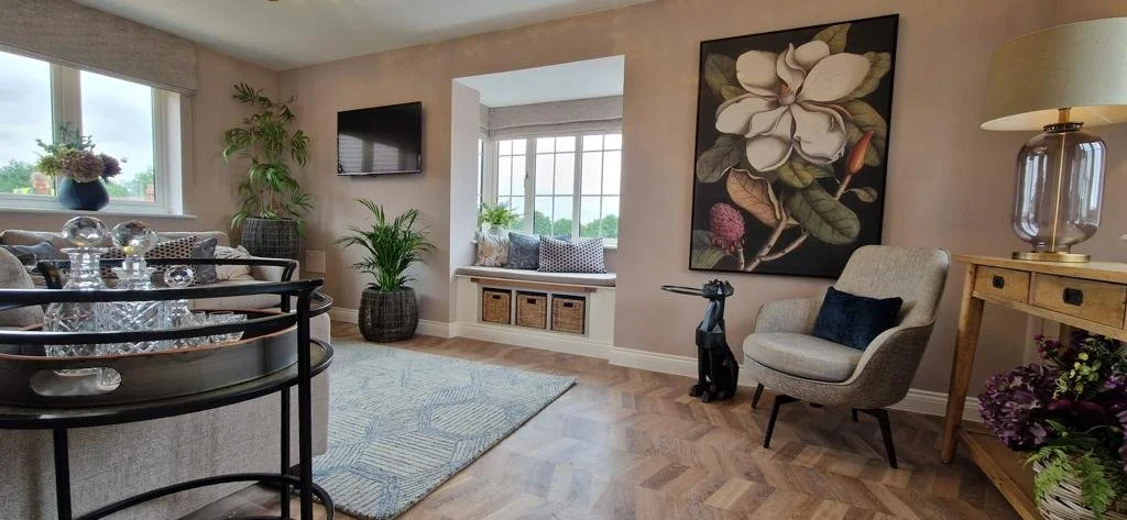 Living room with large window, plants, wall-mounted TV, decorative ottoman, armchair, wooden side table, floral artwork, black dog-shaped sculpture, and a lamp.