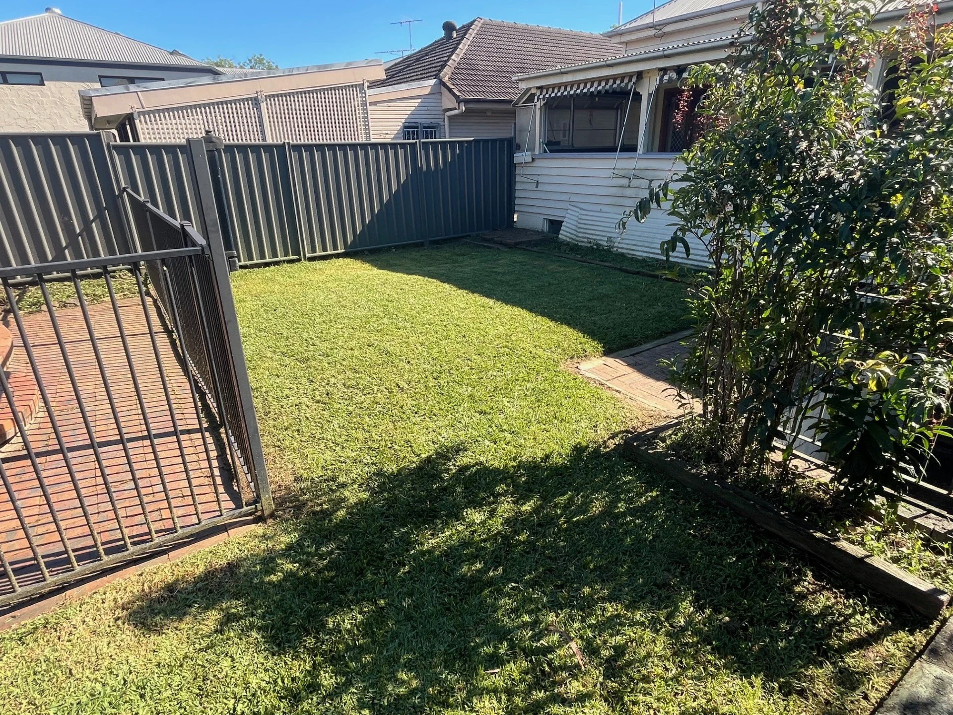 A backyard with green grass, a metal fence, a small brick path, and a white house with a window and awning in the background under a clear blue sky.