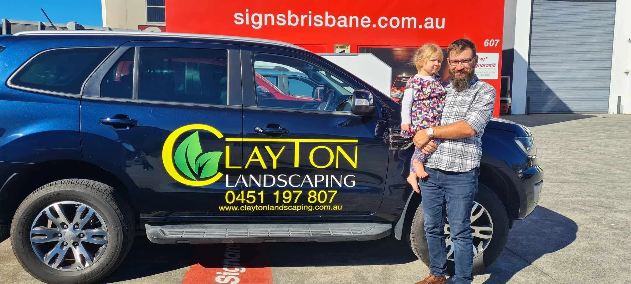 A man holding a young girl stands next to a black Clayton Landscaping vehicle parked outside a building with a red sign in the background. The vehicle has yellow and green branding and contact information for Clayton Landscaping.