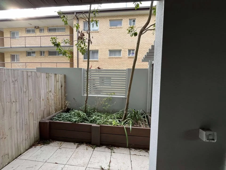 A small backyard garden with three thin trees, green plants, and bushes in a rectangular wooden planter. The yard is fenced with a wooden fence on the left and a white privacy screen at the back. There are tiled paving stones on the ground.
