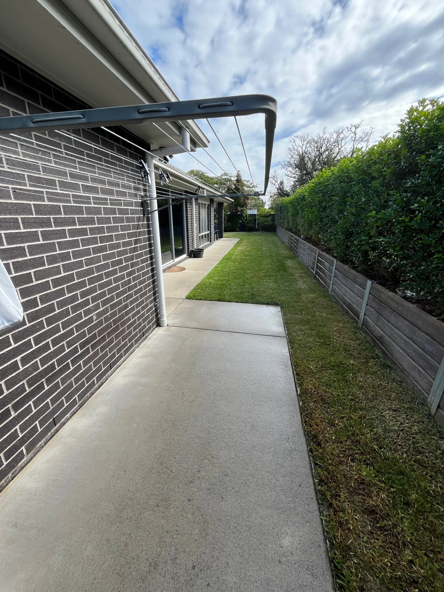 Side yard with concrete patio, patch of grass, a black brick wall, and a wooden fence with bushes on the right. Clothes drying rack is mounted to the house.