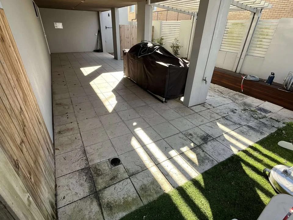 Empty outdoor patio area with a covered BBQ grill, a small garden bed, and a wooden deck, all enclosed by a privacy fence.