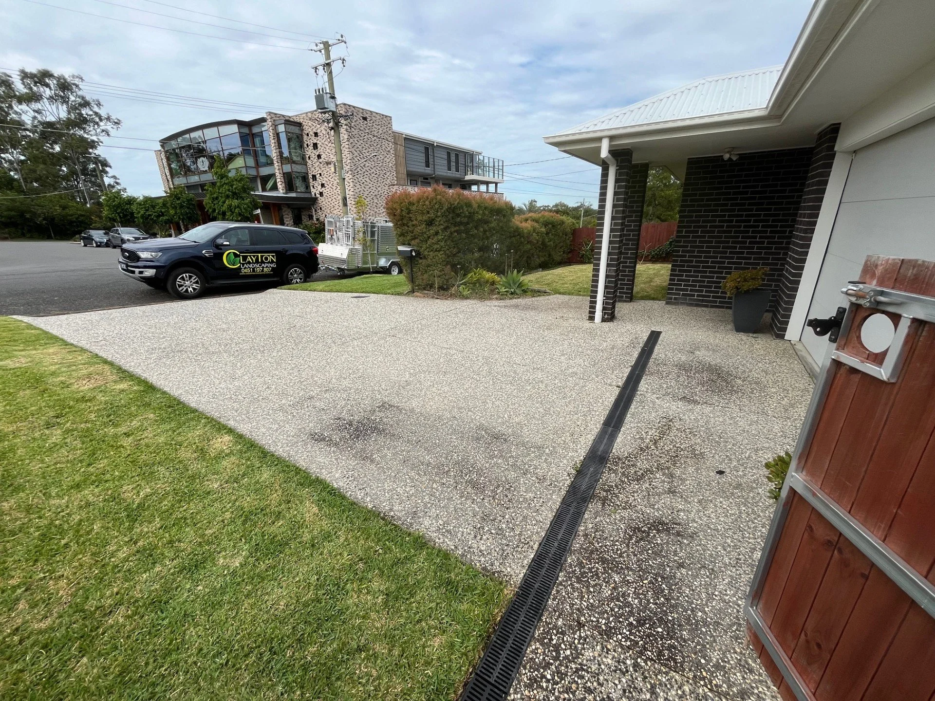 View of a house's driveway with a black landscape company vehicle parked nearby, adjacent to a modern multi-story residential building, with a grassy yard and bushes, on an overcast day.