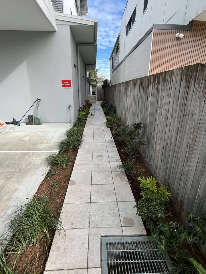 A narrow outdoor walkway with light gray tiles, bordered by a wooden fence on the right and a building with a gray wall on the left, along with plants and garden beds on both sides, and a parking area visible to the left. A fire hydrant sign is on th