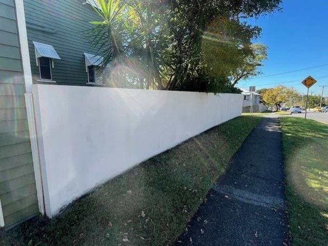 Sidewalk and grassy area next to a white privacy fence with trees and a building in the background under a clear blue sky.