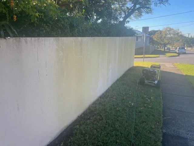 A white exterior wall with some streaks and patches, next to a grassy area and a sidewalk, with a small pressure washer on the lawn.