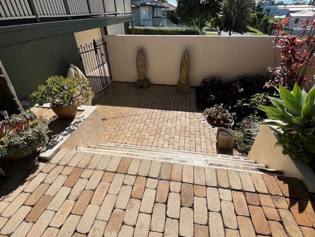 Brick-paved outdoor patio with potted plants and garden statues, enclosed by a white wall and neighboring houses in the background.