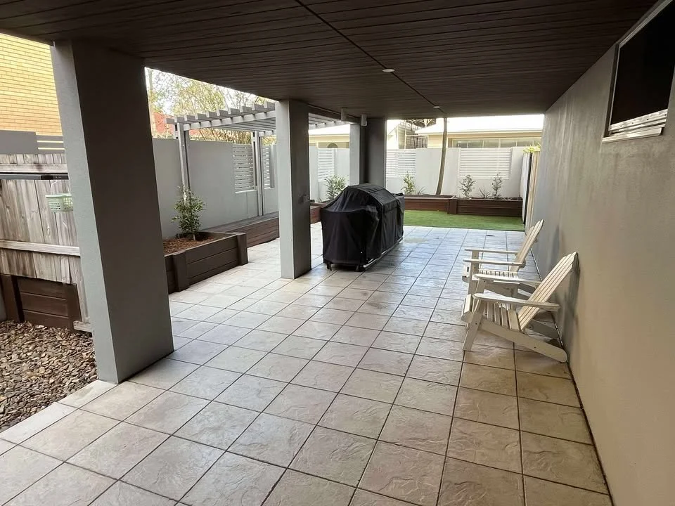Covered backyard patio with tiled floor, two white outdoor chairs, a black covered grill, planter boxes with plants, and a small grassy area in the background.
