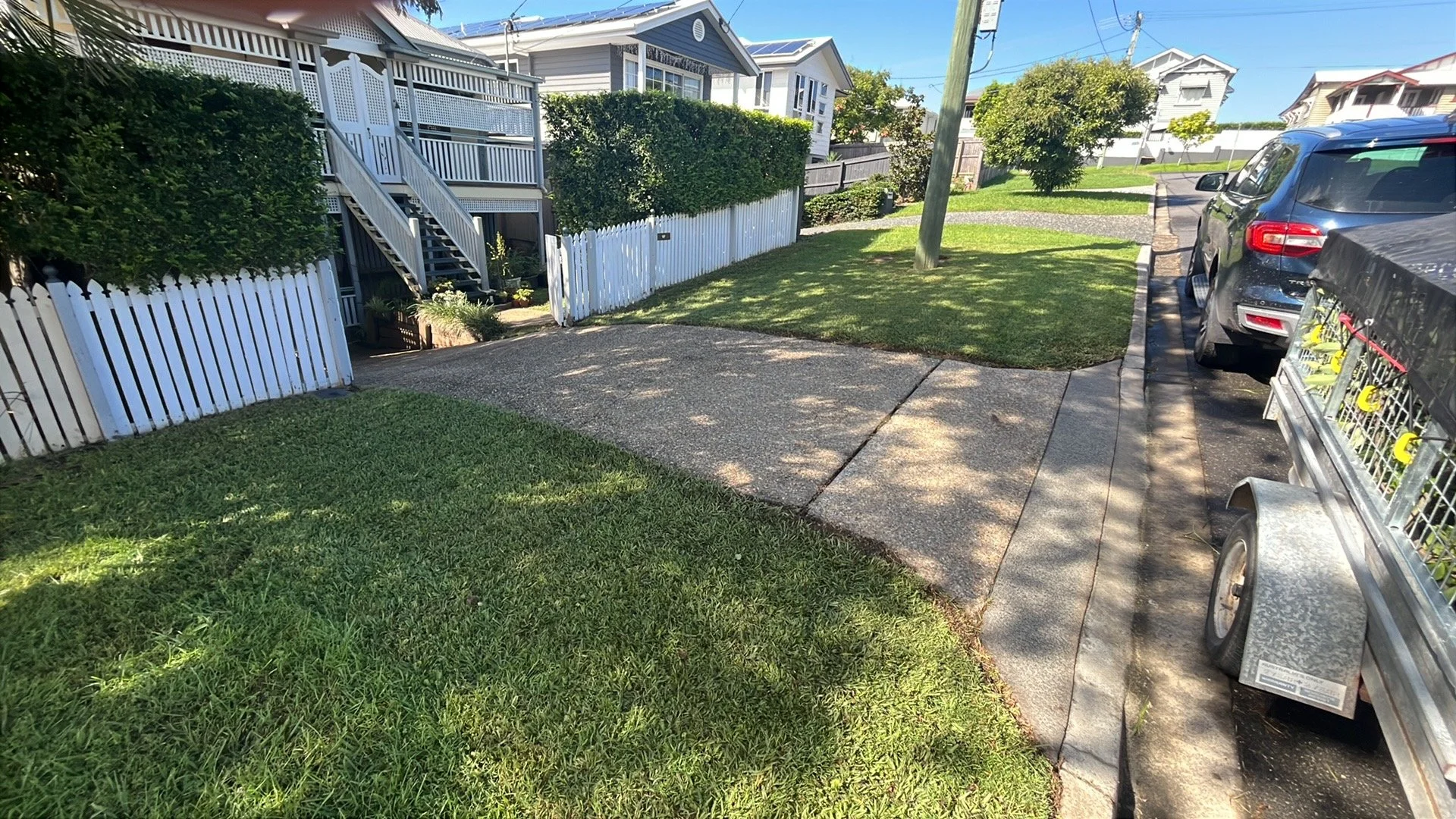 Residential front yard with a concrete driveway, green grass, and a white picket fence. There are houses in the background, parked cars, and a utility pole with power lines.