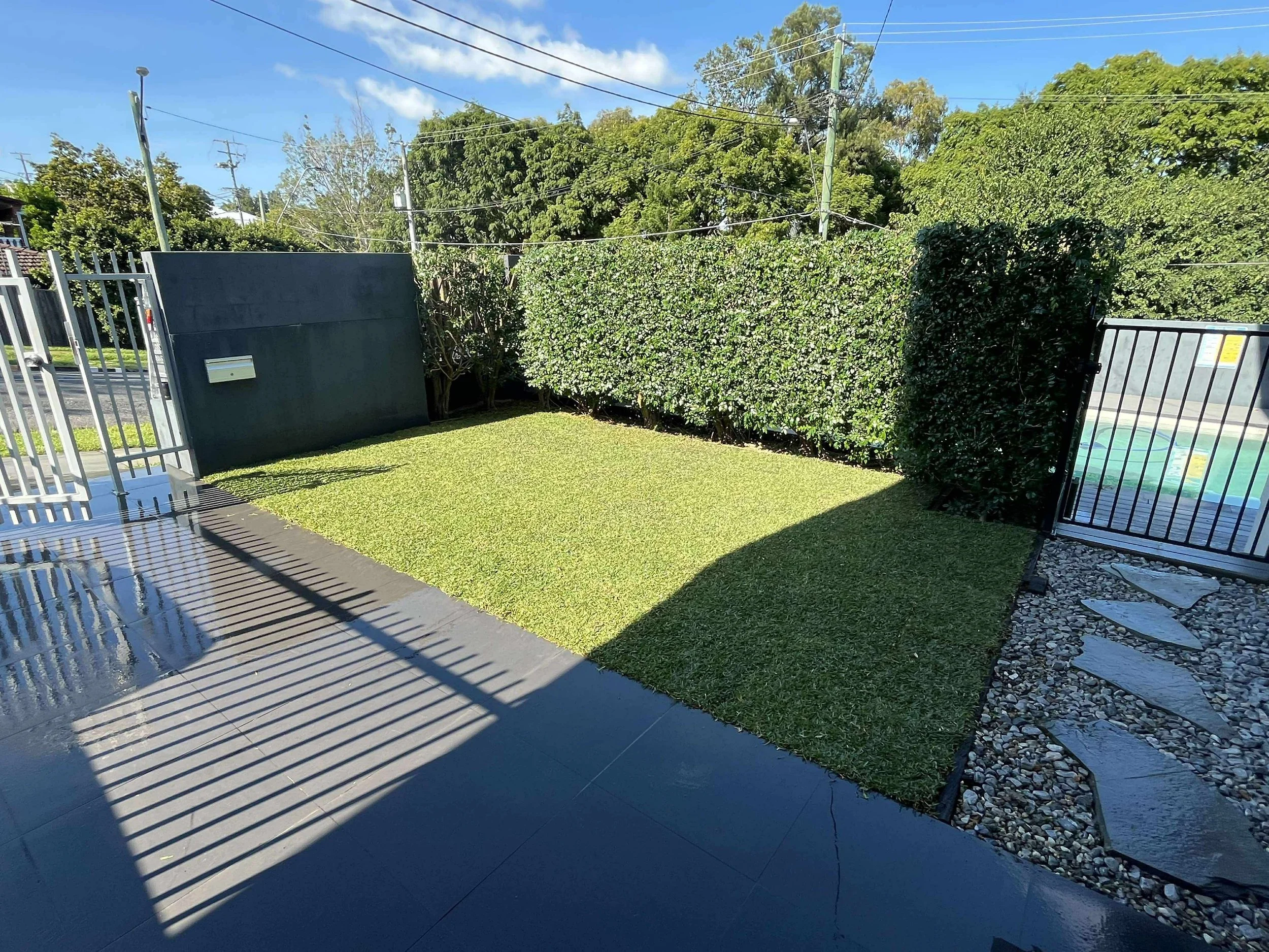 Small backyard with neatly maintained grass, surrounded by tall green hedge, with a sliding gate on the right, a shadow cast on the ground, and a paved walkway with stones on the right side.