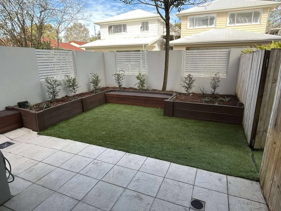 Small backyard with artificial grass, wooden garden beds with plants, a tree in the center, white fence, and neighboring houses visible beyond.
