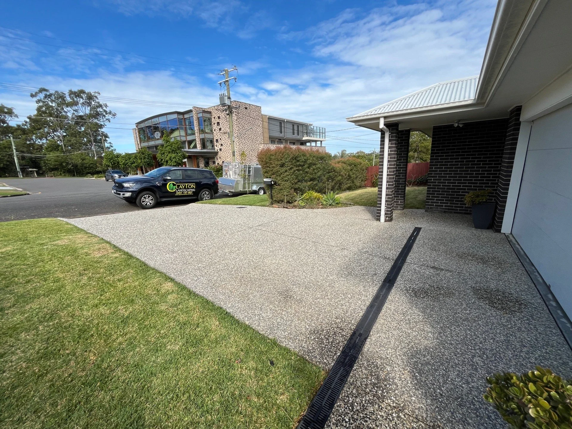 View of a residential driveway bordering a grassy lawn, with a black vehicle parked facing the street, a trailer attached to the vehicle, and a modern multi-story building across the street under a partly cloudy sky.