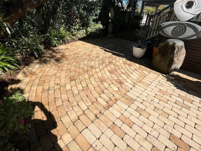 Brick patio with curving pattern, surrounded by plants, a large stone, and outdoor sculpture.