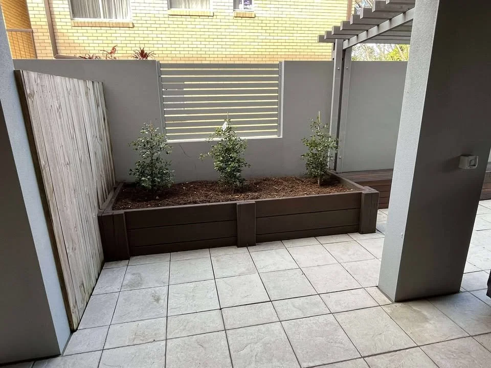 Small backyard patio with white tiles, a wooden planter box with three small plants, a wooden fence on the left, a concrete wall with a small window, and a gray pergola on the right.