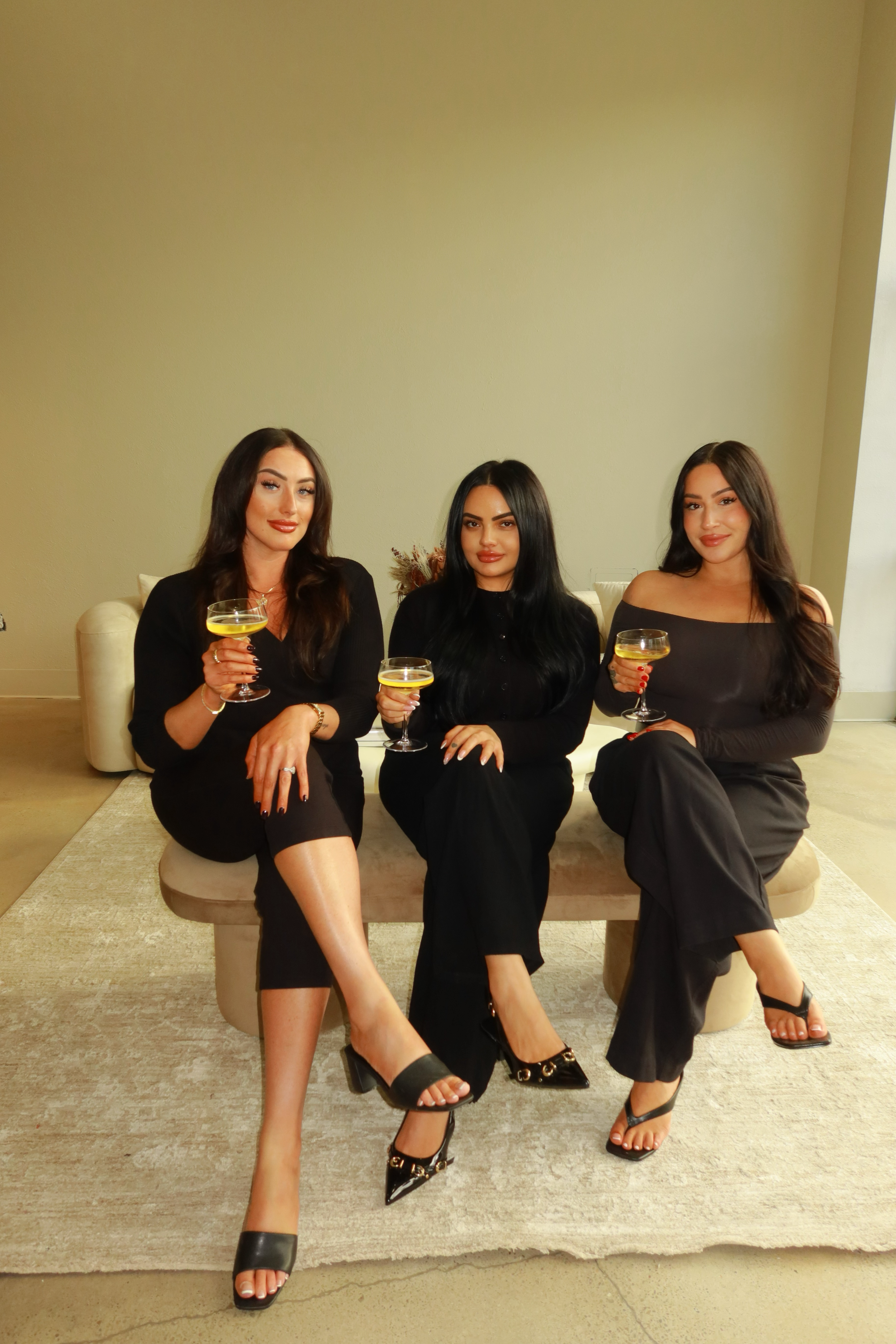Three women dressed in black sitting on a bench, each holding a cocktail glass, in an indoor setting with a plain wall behind them.