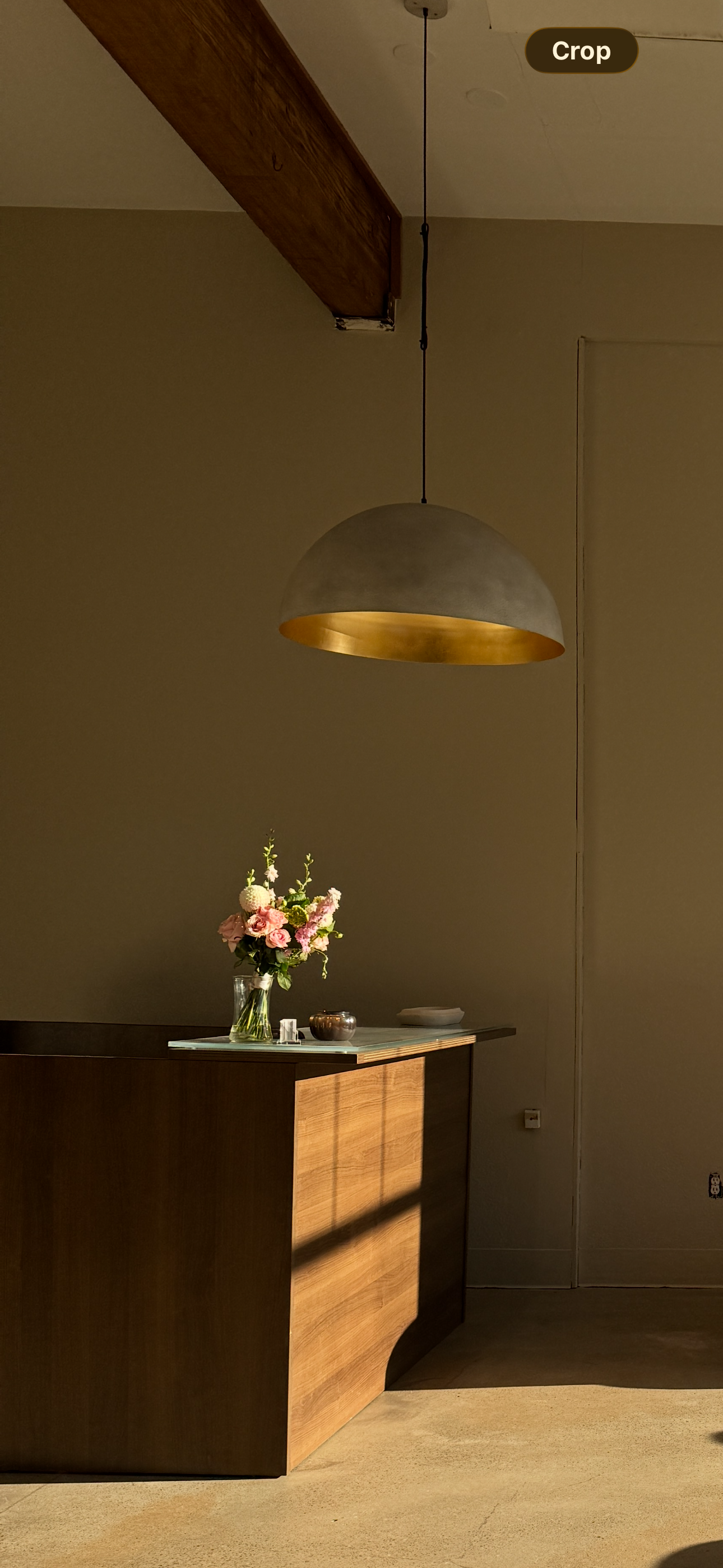A modern interior scene featuring a wooden console table with a vase of pink and white flowers, a small metal bowl, and a white dish. A pendant light with a gold interior hangs from the ceiling. The wall behind is plain and beige.