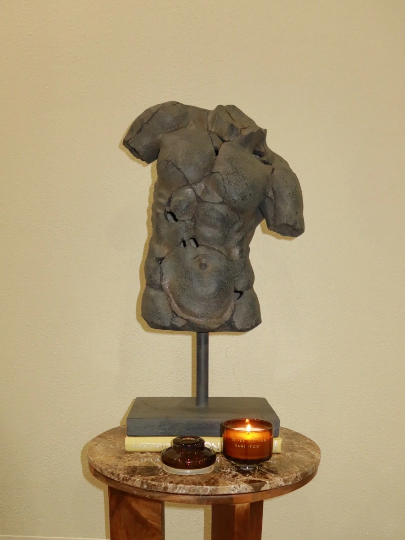 A sculpture of a female torso made of stone cracks, displayed on a stand. It sits on a round marble table with a candle and a small jar in front.