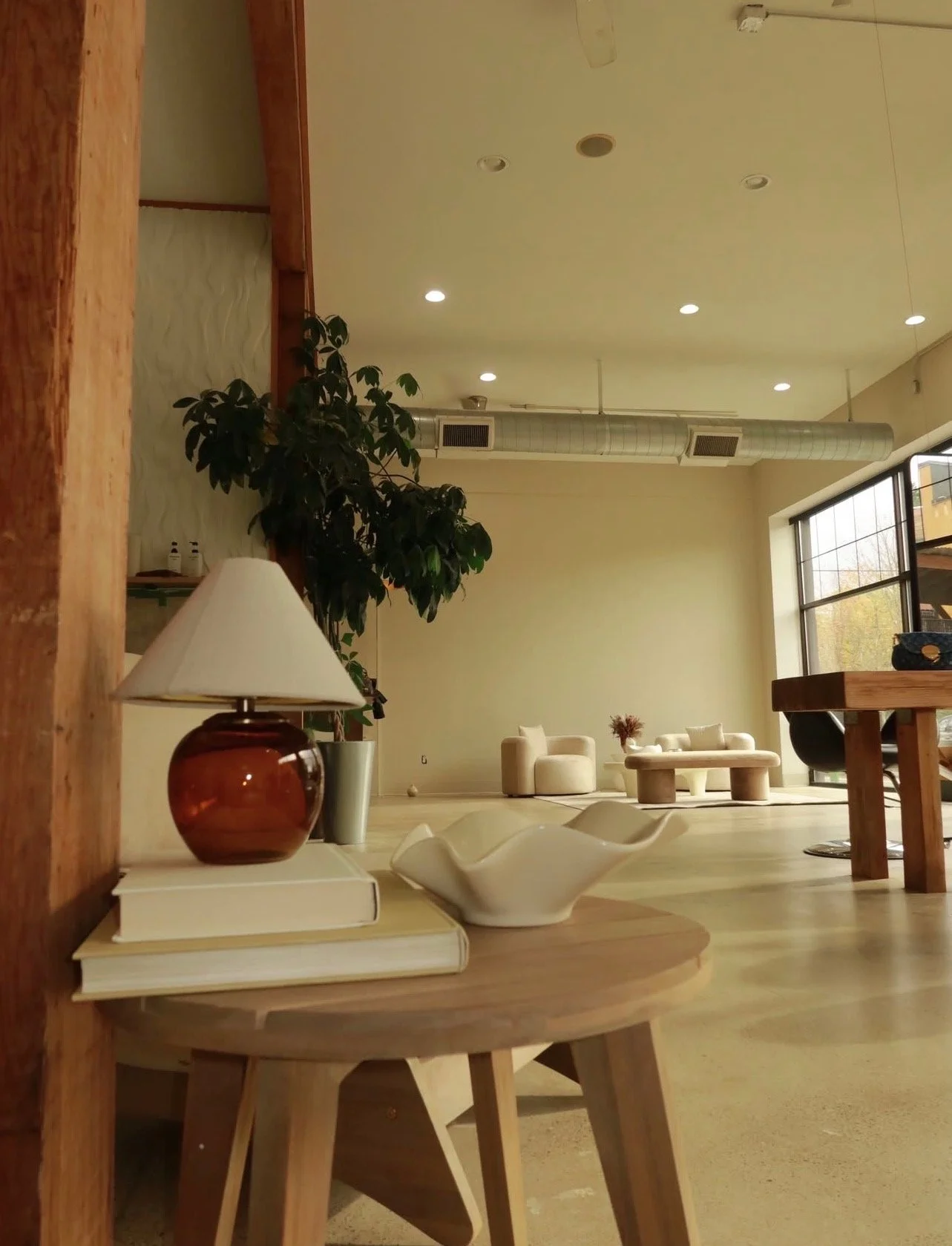 Interior of a modern, minimalist lounge with cream-colored furniture, large window, indoor plants, and decorative items like a lamp and ceramic dish
