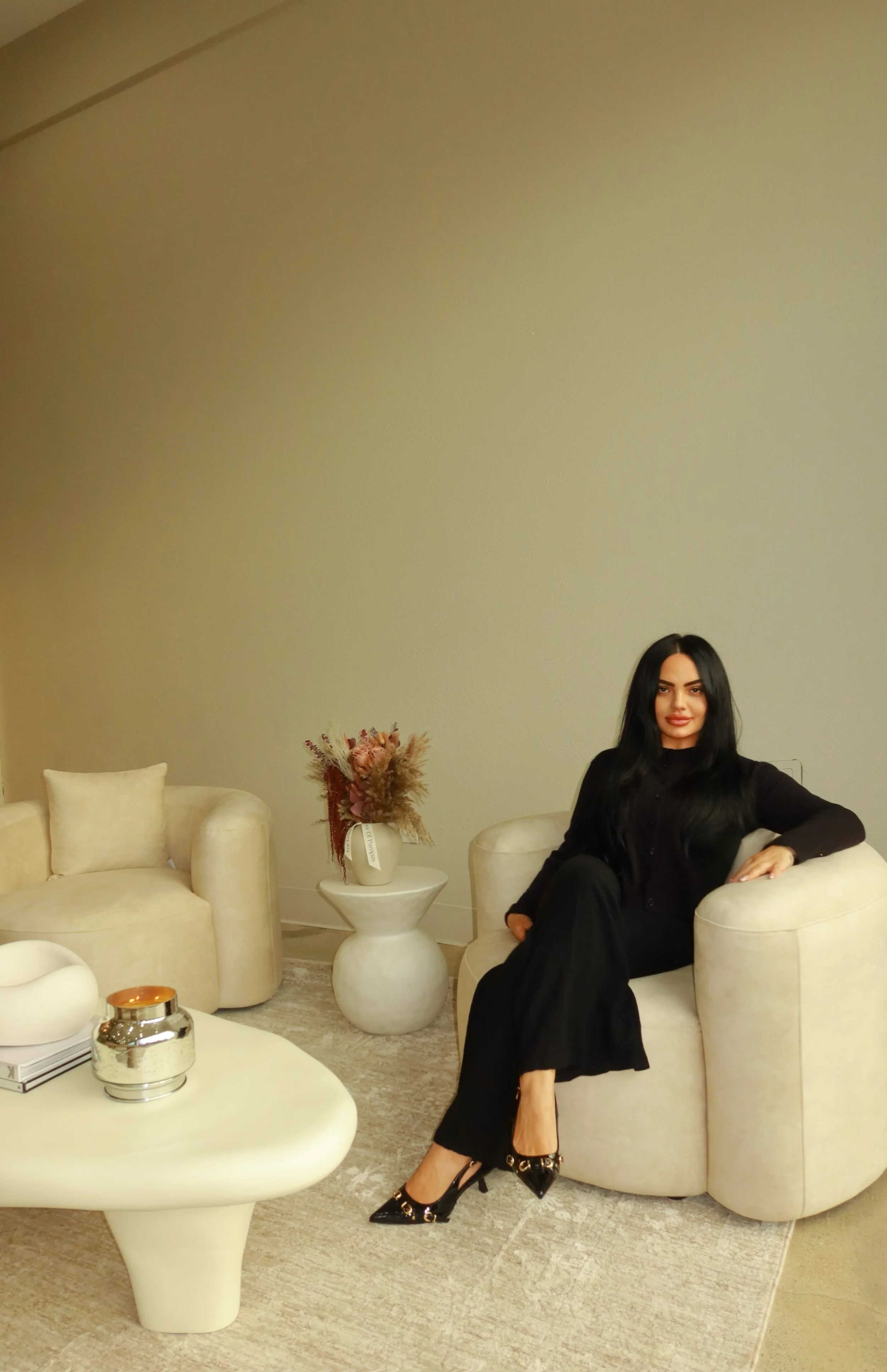 A woman with long dark hair, wearing a black outfit and black heels with gold embellishments, is sitting on a beige armchair in a stylish, modern living room. The room features a white coffee table with decorative items, a beige sofa with pillows, a small white side table with a vase of dried flowers, and a large light-colored wall.