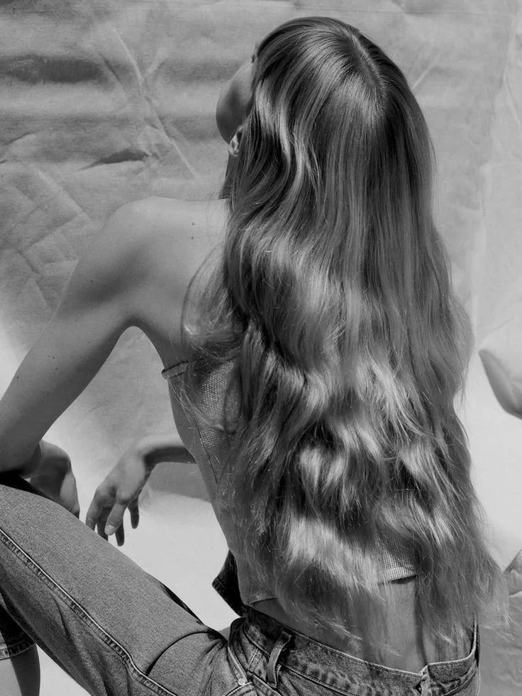 A woman with long, wavy hair sitting on a chair with her back to the camera, wearing jeans and a sleeveless top.