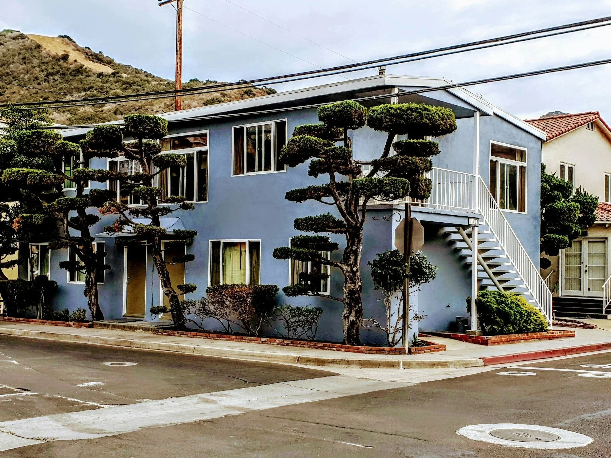A blue two-story building with white staircases and trim, surrounded by neatly sculpted trees and shrubs, in an urban area with parking spaces in the foreground and hilly terrain in the background.