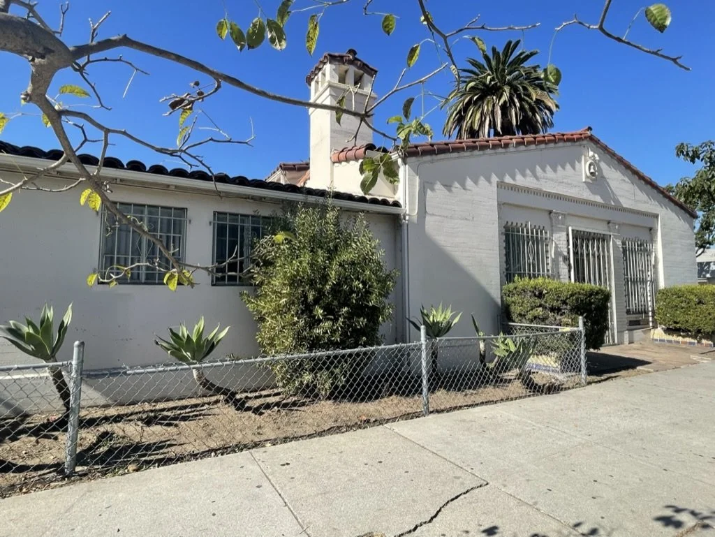 A white house with barred windows and a red-tiled roof, surrounded by a small garden with various plants and a chain-link fence, under a clear blue sky.