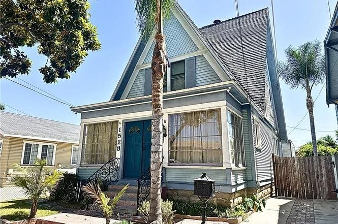 A blue A-frame house with large front windows and a staircase leading to the front door, surrounded by palm trees and a garden.